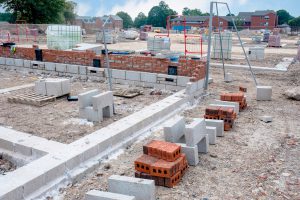 Canteiro de obras com tijolos maciços e blocos de cimento em destaque. Ao fundo, construções de alvenaria estrutural.