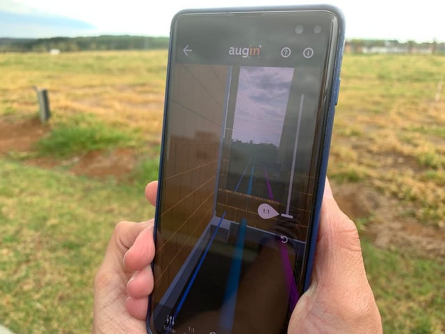 Uma foto da mão segurando o celular e nele está visualizando uma projeção virtual de uma construção no terreno real.