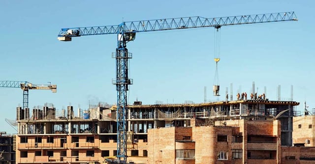 Um canteiro de obras e um edifício ainda sem acabamento. Trabalhadores e guindastes são vistos junto da construção