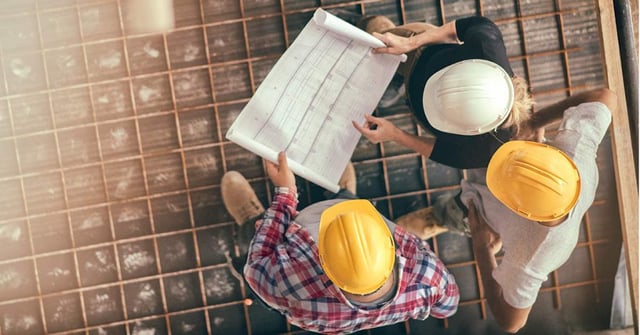 Uma foto de três pessoas vistas de cima, em uma discussão de trabalho. Estão usando capacetes em um canteiro de obras