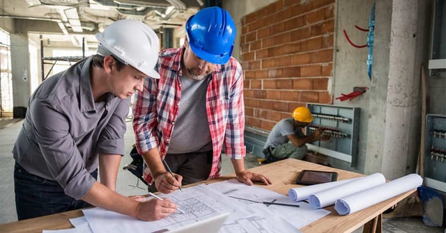 Dois homens em uma construção examinando planos arquitetônicos, ambos estão usando capacetes, ao fundo, um terceiro homem