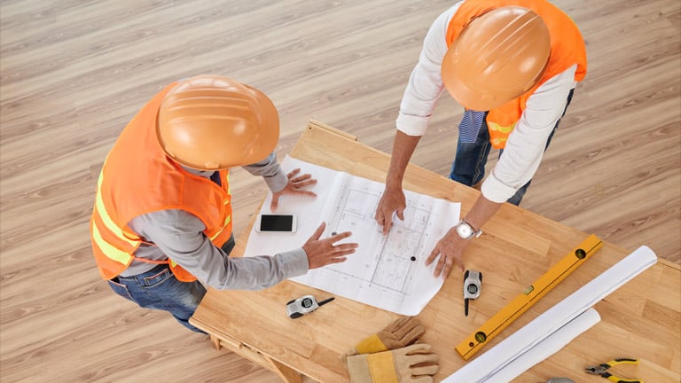 Dois trabalhadores de construção debruçados sobre a mesa examinando planos arquitetônicos. À mesa tem ferramentas de trabalho