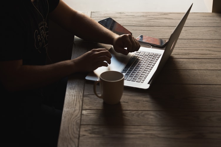 A foto foca nas mãos de uma pessoa sentada à mesa trabalhando em um laptop. Há uma caneca, um smartphone.